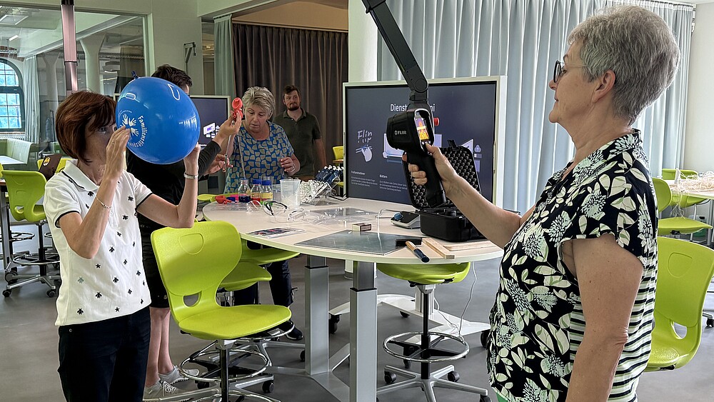 Foto: HSZG Zwei Lehrerinnen testen Versuche im School Lab. Eine hält einen blauen Luftballon vor ihr Gesicht, die andere hat ein digitales Messgerät in der Hand.