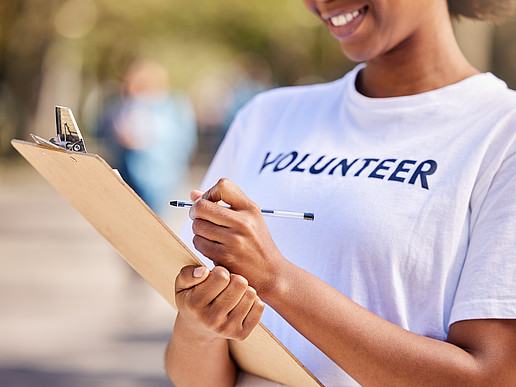 Eine dunkelhäutige junge Frau im weißen T-Shirt mit der Aufschrift "Volunteer" hält einen Stift und eine Kladde in den Händen und trägt lächelnd etwas darauf ein. 