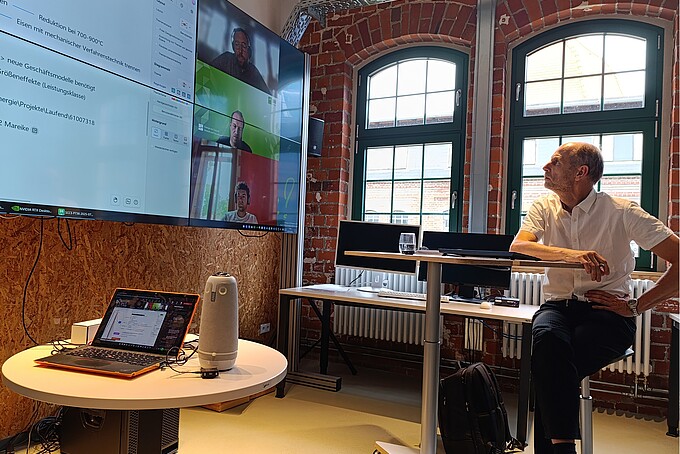 Professor Zschunke sits on a high chair and looks at a large video screen in a workroom with brick walls and semicircular windows.