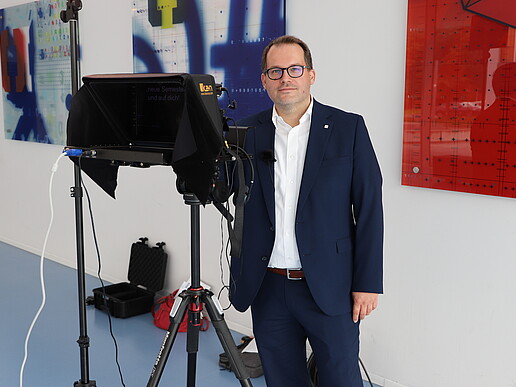 The principal stands in the foyer of house four next to a teleprompter and smiles into the camera.