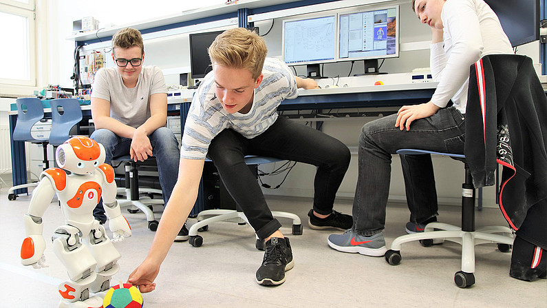 Drei Schüler in einem Labor mit einem Roboter und einem Ball