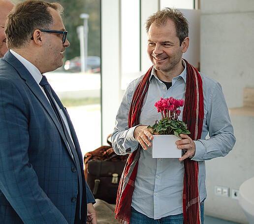 The rector is talking to a professor who is holding a potted plant in his hands.