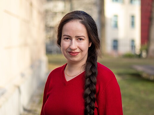 Anna Prenzel smiles into the camera, standing on a meadow next to a campus building in Görlitz and wearing a red top.