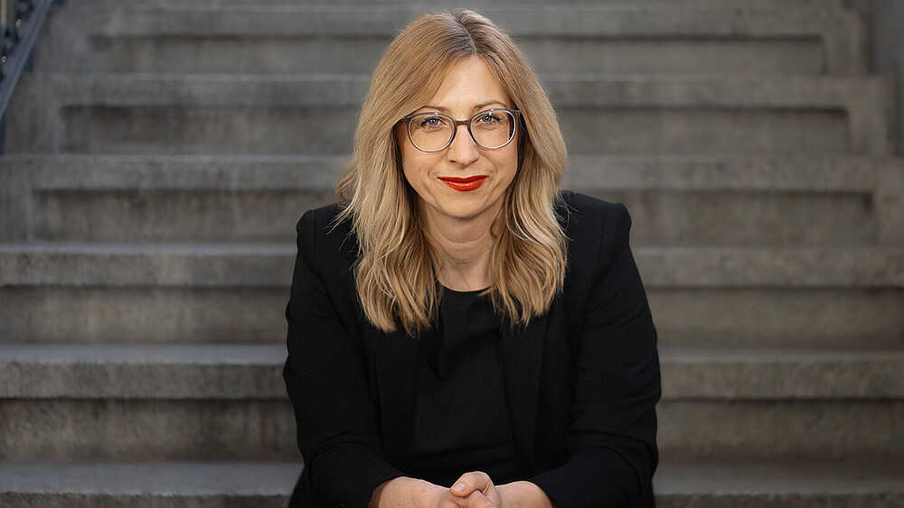 Prof. Dr. rer. pol. Jana Stolz-Römmermann wears a black blazer and glasses, sits on a step and smiles into the camera.