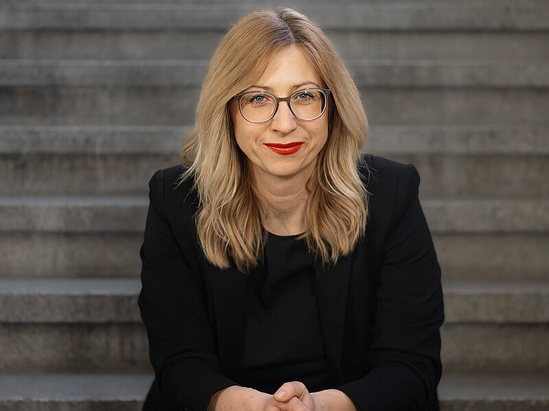 Prof. Dr. rer. pol. Jana Stolz-Römmermann wears a black blazer and glasses, sits on a step and smiles into the camera.