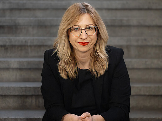 Prof. Dr. rer. pol. Jana Stolz-Römmermann wears a black blazer and glasses, sits on a step and smiles into the camera.