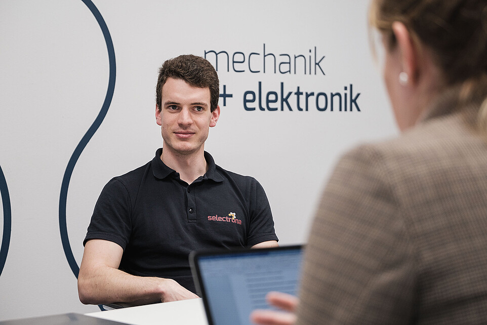 Man in a black polo shirt sits at a table and listens.