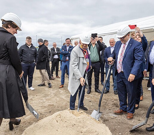 The principal holds a shovel in his hand and picks up a shovelful of sand. Next to him, the other project team members do the same. Guests of the event stand in the background.