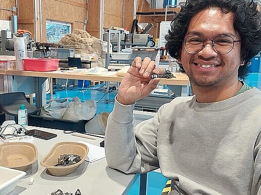 Faiz sits at his workstation in the Lander Technikum. In front of him are black plastic parts.
