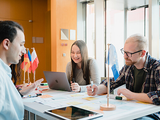 Students are sitting at their desks working on a project. In front of them is a poster with sticky notes, next to it are tablets and notebooks. The German, Polish and Czech flags are on the table.