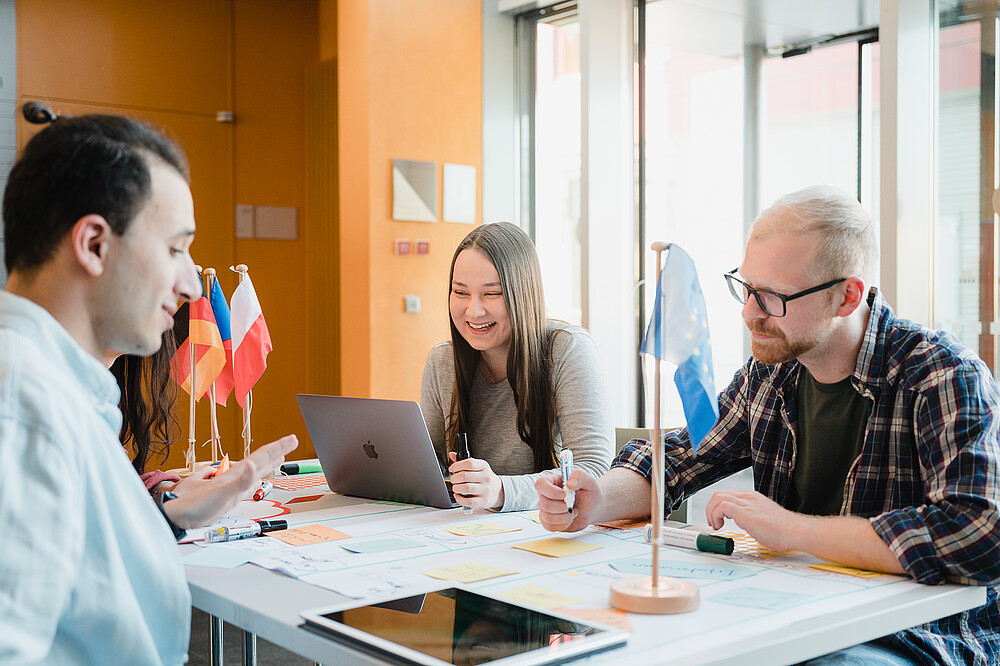 Students are sitting at their desks working on a project. In front of them is a poster with sticky notes, next to it are tablets and notebooks. The German, Polish and Czech flags are on the table.