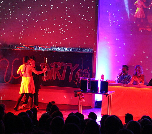 A dance couple dances on stage in the red light and is watched by the jury from the judges' table.