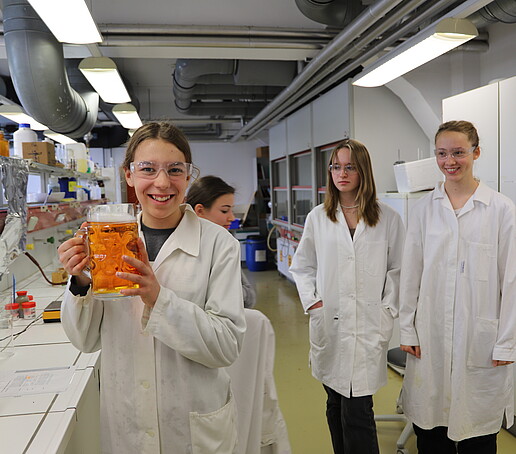 A schoolgirl holds a beer mug with an orange liquid with white foam in her hand and smiles at the camera.