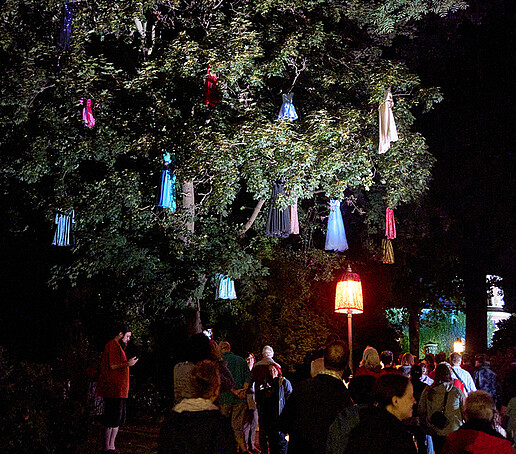 Colorful cloths hang in a tree, visitors walk underneath.