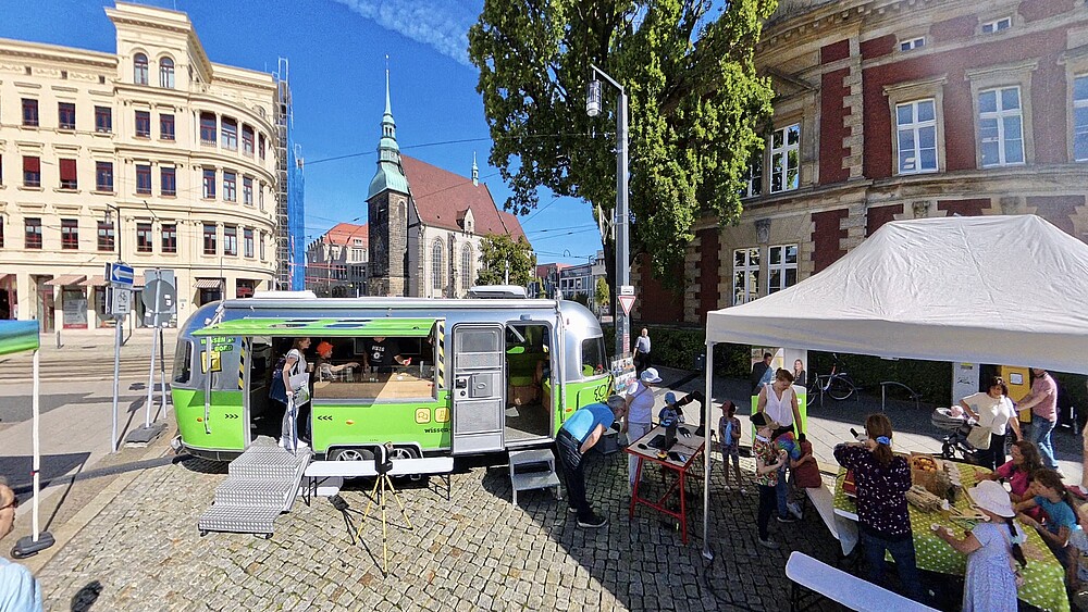 Foto: Aileen Baumgärtner Der Mobile Lernort-Anhänger steht mit offenen Türen und Seitenfenster auf dem Görlitzer Postplatz. Daneben steht ein weißer Pavillon. Darunter beschäftigen sich Kinder und andere Gäste mit den ausgelegten Materialien.