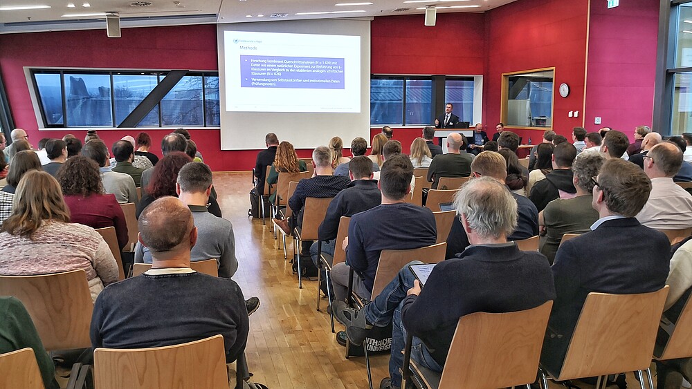 In einem gut gefüllten Seminarsaal mit roten Wänden hält ein Mann einen Vortrag neben einer Leinwand, auf der die Präsentation abgebildet ist.