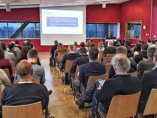 In a packed seminar room with red walls, a man gives a lecture next to a screen showing the presentation.