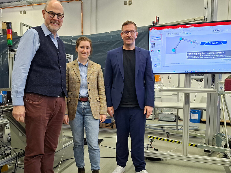Three people are standing in the laboratory, next to a screen on which a graphic can be seen.