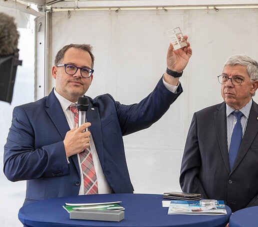 The Rector of the HSZG holds a metal HSZG signet in his hand at a lectern in a tent. A man stands next to him.