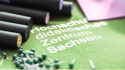 Ein grüner Ausdruck mit der Aufschrift Hochschuldidaktisches Zentrum Sachsen liegt auf einem Tisch umgeben von Stecknadeln. 
