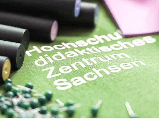 A green printout with the inscription Hochschuldidaktisches Zentrum Sachsen lies on a table surrounded by pins.