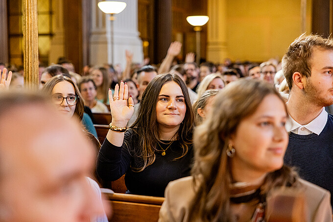 A young woman is sitting in a church pew among other guests and raises her hand.