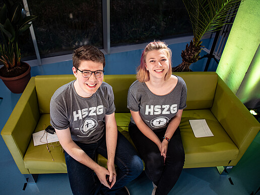 Cornelius Baier and Carolin Renner sit on the green presentation sofa and look happily up into the camera