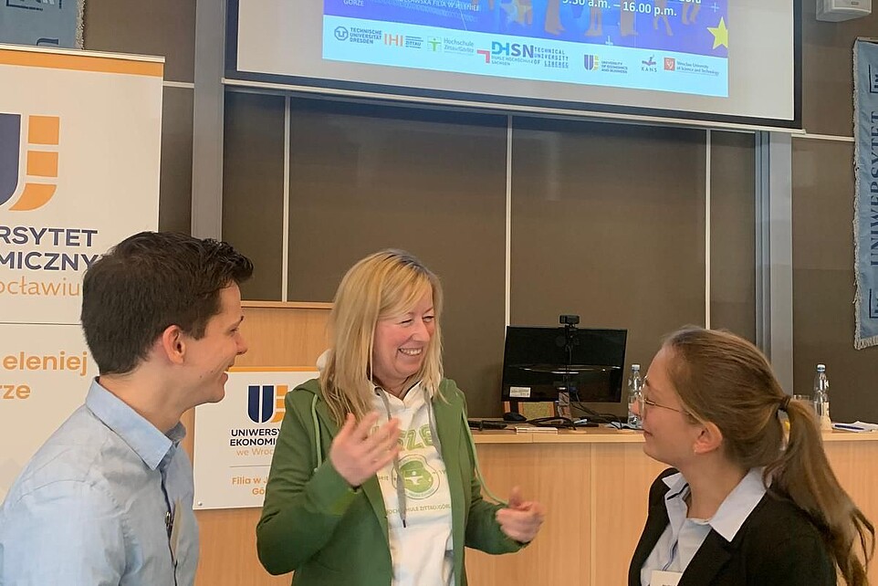 The Vice President in conversation with two young researchers from the HSZG at the conference. A presentation lights up on a screen in the background.