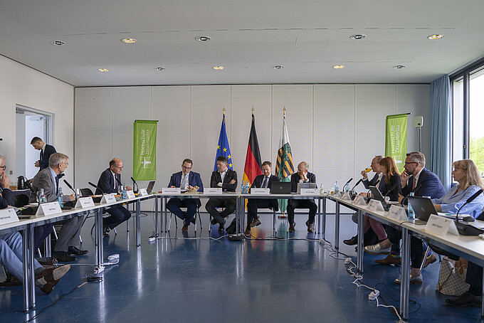 Beteiligte der Kabinettssitzung sitzen in einem Halbkreis mit Bannern der 'Hochschule' im Hintergrund.