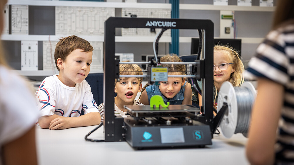 Four children are sitting in the electrical engineering laboratory looking at a small green elephant on the platform of a device.