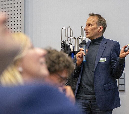 A lecturer speaks into a hand-held microphone in front of the audience.