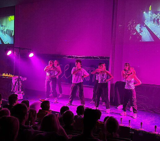 A dance group dances on the podium in the purple light.