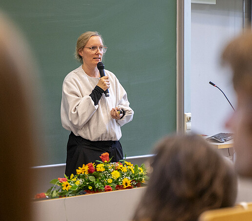 A woman speaks into a microphone in front of a green board in the lecture hall.