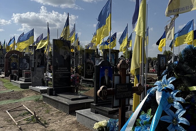 Ein Soldatenfriedhof ist übersät mit ukrainischen Flaggen, die im Wind wehen. Auf den Grabsteinen sind Porträts, Namen und Daten eingraviert. Blumen und Kränze schmücken die Gräber, während der blaue Himmel mit einigen Wolken einen friedlichen Kontrast zur ernsten Szenerie bildet.