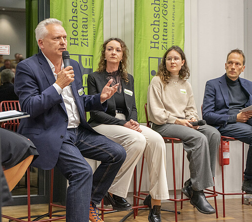 In the panel, four guests sit next to each other on high chairs, one guest speaks into a microphone.