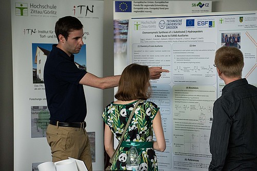 Ein Forschungsmitarbeiter zeigt zwei Besuchern an der Posterwand seine Forschung