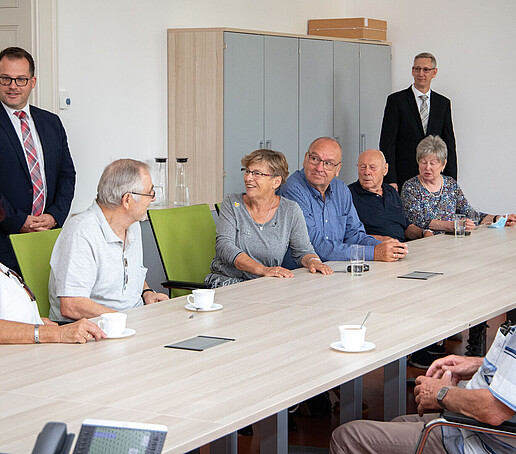 Ältere ehemalige Studierende sitzen an einer Tafel im Senatssaal. Der Rektor und Prof. Bellair stehen im Hintergrund und unterhalten sich mit ihnen.