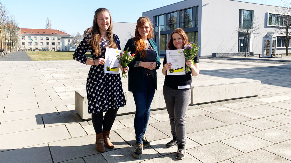 Zwei Studentinnen und die Moderatorin vom HIT stehen auf der Campusachse vor Haus Z 4. Die Studentinnen halten ihre Urkunden in die Kamera und lächeln.