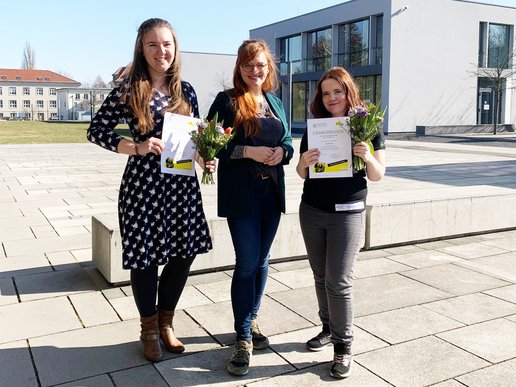 Two female students and the moderator from HIT stand on the campus axis in front of House Z 4. The students hold their certificates up to the camera and smile.