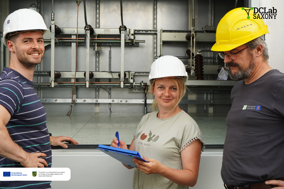 Two employees, one of whom is wearing a helmet, are standing in front of a test facility