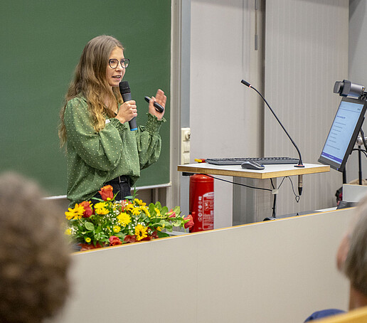 A young scientist speaks into a microphone in front of a green board in the lecture hall.
