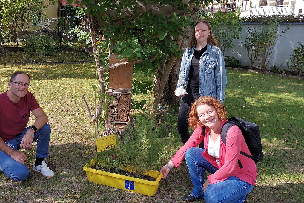 Im Garten lachen ein Professor, seine Tochter und eine Hochschulmitarbeiterin in die Kamera. Sie stehen neben einem selbstgebauten Insektenhotel.