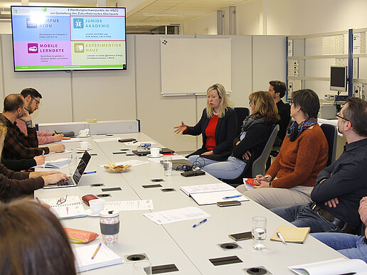 Teachers sit at a long table, a screen hangs in the background.