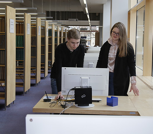Zwei Auszubildende beugen sich in der Bibliothek über einen Rechner.