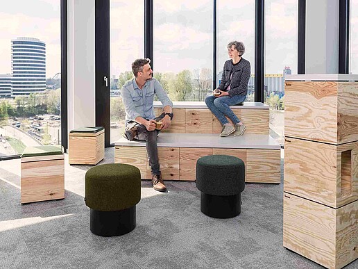 An advertising photo of two people talking while sitting on modern furniture in an office building with large windows in the background.