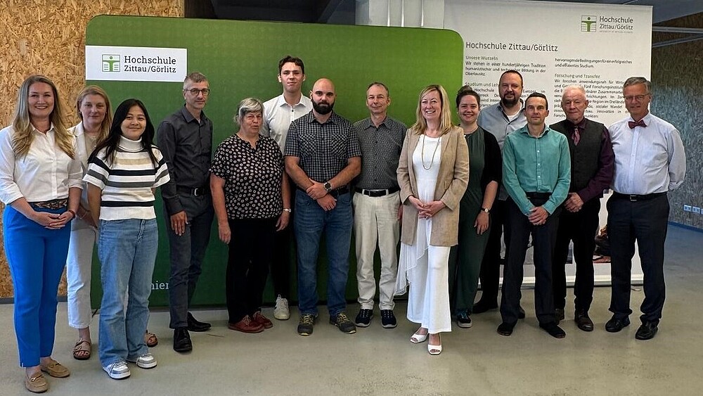 Representatives of the HSZG and Infineon Technologies stand next to each other in front of a green press wall and a HSZG roll-up in the Celsiuz for a group photo.