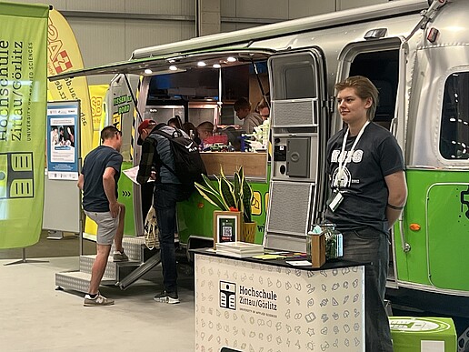 The Airstream trailer stands in the exhibition hall and visitors enter it to look at experiments. In the foreground is an employee at an HSZG counter.
