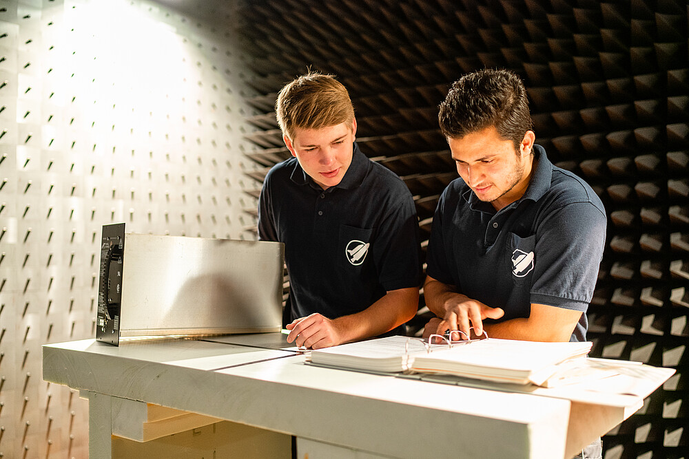 Zwei KIA-Studenten blättern in einem offenen Ordner und stehen im Energietechniklabor.