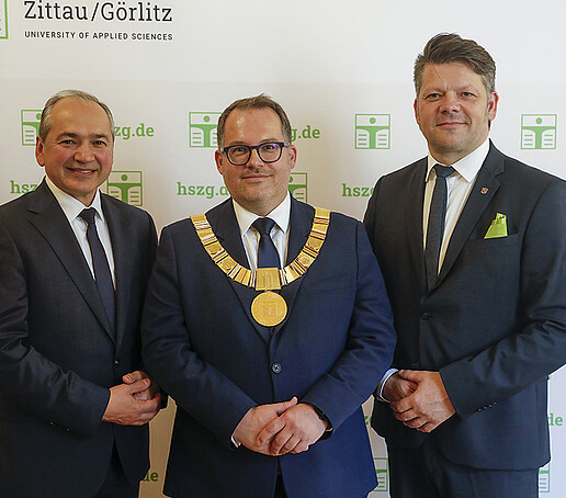 Mr. Ursu, Prof. Kratzsch and Mr. Zenker stand next to each other in front of a press wall and look into the camera.