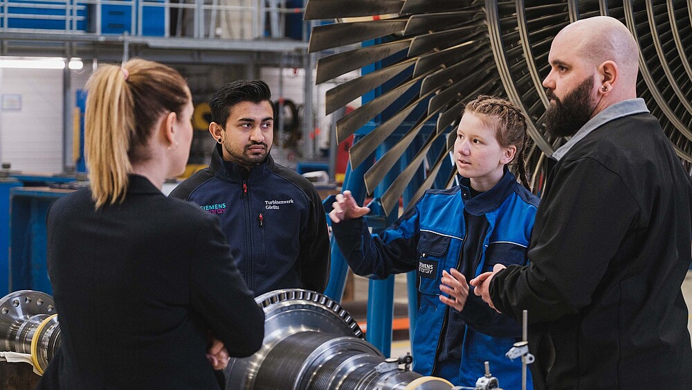 Sandeep und Alina stehen in einer Werkshalle vor einer Turbine und unterhalten sich mit der KIA-Koordinatorin der HSZG und einem Siemens-Mitarbeiter.